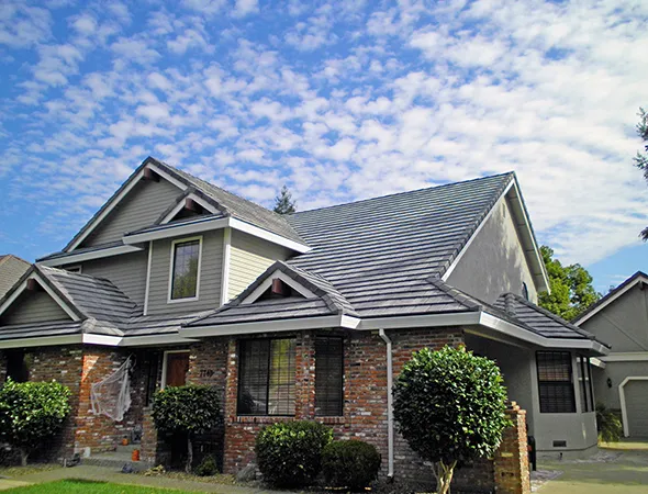 A gray house stands under a clear blue sky, surrounded by trees, showcasing its well-maintained roofing and serene environment.