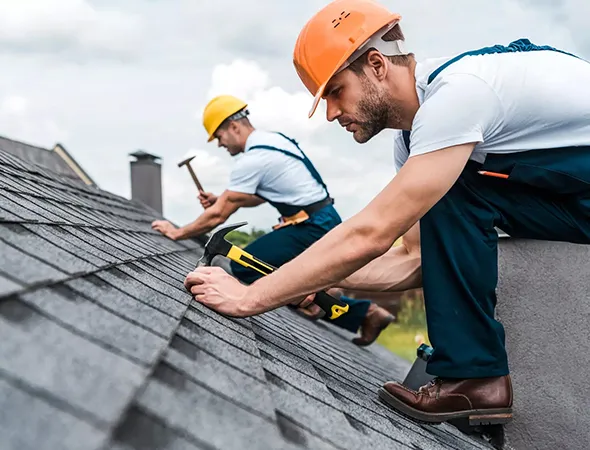 Two men are engaged in roofing work, using hammers to secure materials on a sloped roof under a clear blue sky.